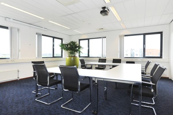 Vergaderruimte aan de Laan van S-Gravenmade met moderne stoelen, witte tafels in U-opstelling en een grote plant in het midden.